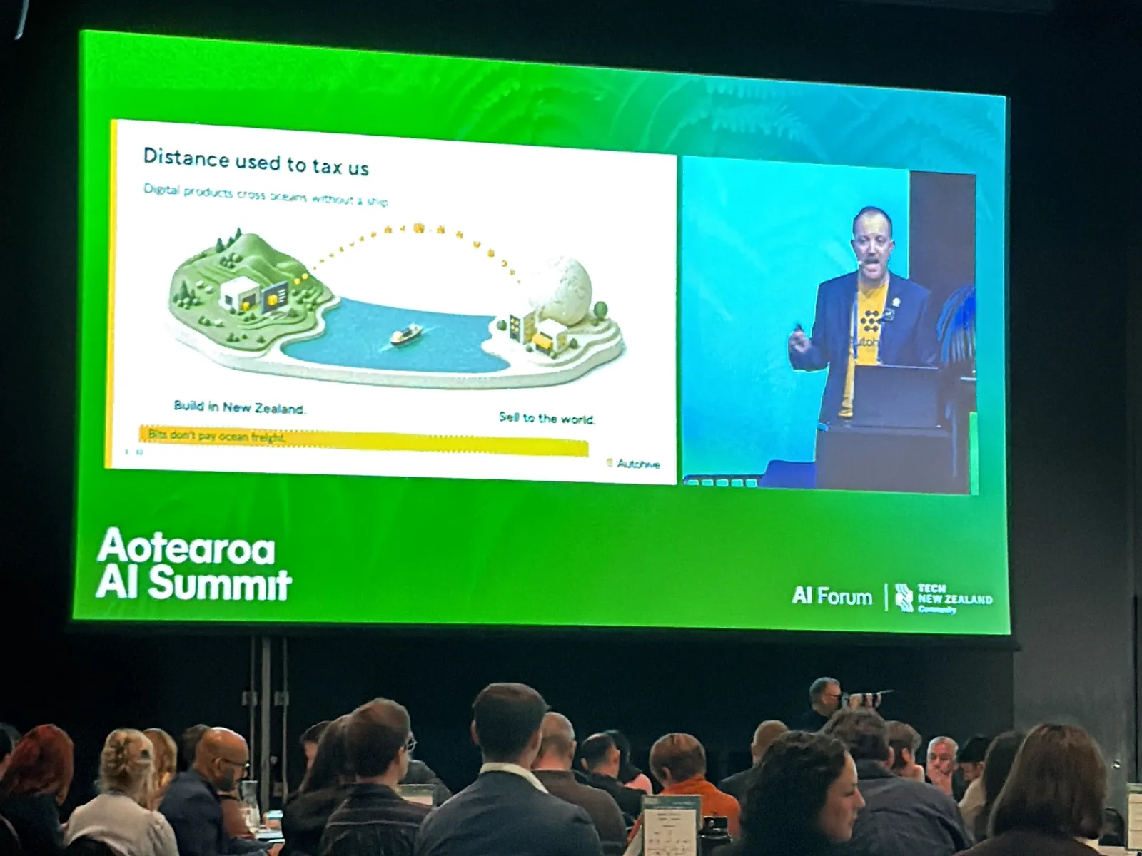 Delegates seated at round tables in the main hall of the Aotearoa AI Summit at Tākina, watching a keynote on a large green summit screen