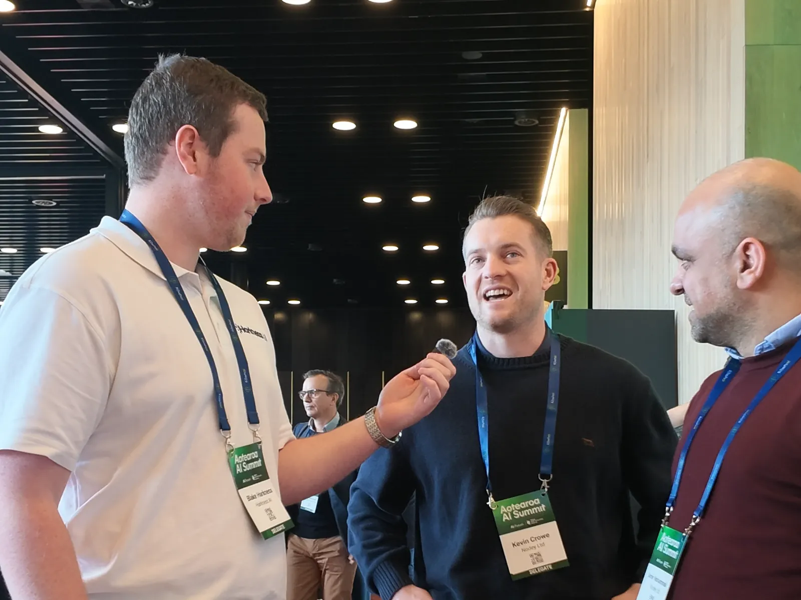 Kevin Crowe laughing as Blake Harkness holds out a microphone, with Amir Mohammadi beside them in the summit foyer