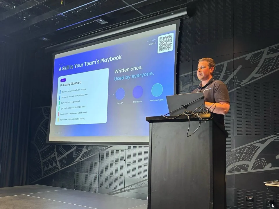 Blake Harkness at the lectern on the Tūranga stage, presenting the slide A Skill Is Your Team's Playbook to business analysts at the BA Big Day In 2026