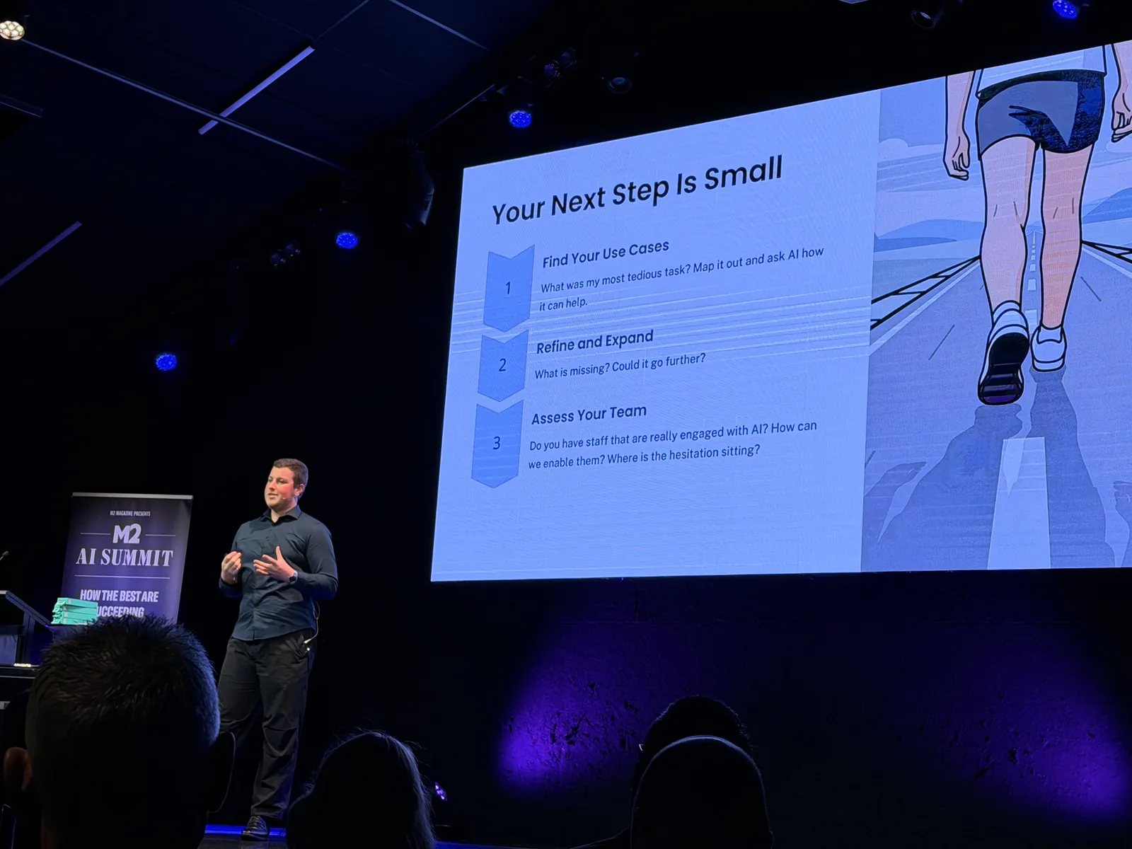 Blake Harkness on stage at the M2 AI Summit in Christchurch, presenting a slide titled Your Next Step Is Small